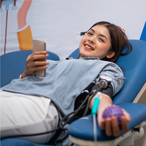 A student smiles while donating blood at the Bleed Blue Blood Drive, holding a stress ball in one hand and a smartphone in the other, lying comfortably on a donation chair.